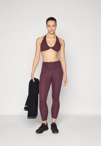Maroon sports bra and high-waisted leggings made of stretchy material. Model holds a black towel. Black athletic shoes complete the look.