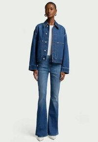 Chaqueta de mezclilla en azul oscuro con un corte amplio, con grandes bolsillos y botones metálicos. Combinada con jeans acampanados azules y calzado blanco.