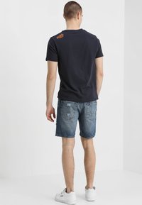 Námořnické bavlněné tričko s oranžovým logem na horní části zad, spárované s poškozenými denimovými šortkami a bílými teniskami, viděné zezadu.