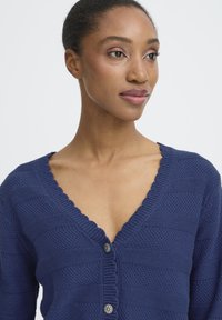 Marineblå strikket cardigan med V-udskæring, der har ribbet tekstur, flæsekanter og dekorative sølvknapper foran.