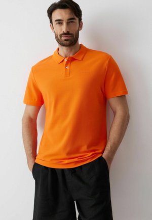 Mężczyzna z ciemnymi włosami i brodą, ubrany w jasnopomarańczową koszulkę polo z krótkim rękawem i czarne spodnie, stojący z rękami w kieszeniach na tle białej ściany.