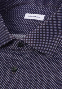 Chemise violet foncé pliée avec un petit motif carré bleu clair et des boutons noirs, avec une étiquette blanche visible contenant le texte "seidensticker".