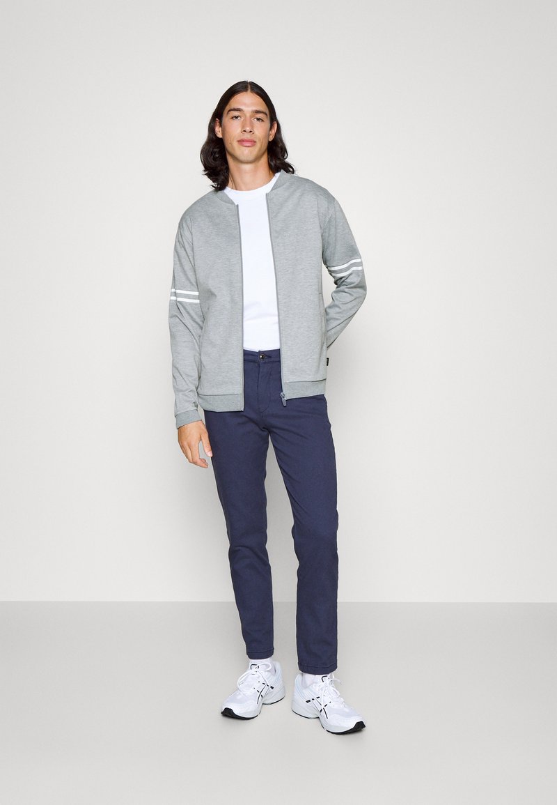 Chaqueta de cremallera gris claro con rayas blancas, combinada con pantalones azul marino y zapatillas deportivas blancas. Textura suave, diseño casual.