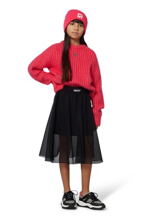 Rode gebreide trui, zwarte doorschijnende mesh rok met logo tailleband en zwarte sport schoenen met roze accenten. Geplaatst tegen een effen achtergrond.