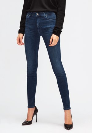 Mujer que lleva vaqueros pitillo azul oscuro, camiseta negra de manga larga y zapatos negros de tacón alto y punta puntiaguda, de pie sobre un fondo blanco.