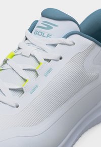 Λευκό παπούτσι γκολφ με εσωτερική επένδυση σε ανοιχτό γαλάζιο, λευκά κορδόνια και νεονί κίτρινες λεπτομέρειες στο πλέγμα κοντά στις οπές των κορδονιών.