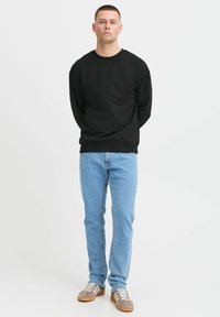 Sudadera negra de manga larga con cuello redondo, combinada con jeans de color azul claro y zapatillas grises con rayas blancas, de pie contra un fondo blanco.