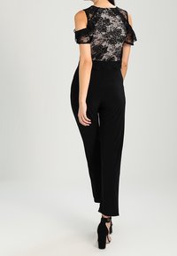 Czarny kombinezon z przezroczystą koronkową górą w kwiatowe wzory, z odsłoniętymi ramionami i długimi, dopasowanymi spodniami; noszony z wysokimi obcasami.