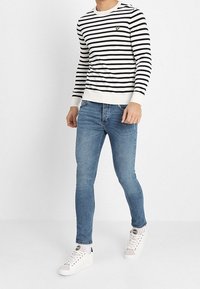 Jersey de manga larga a rayas en blanco y azul marino, con un pequeño logo en el pecho. Combinado con unos vaqueros ajustados de mezclilla azul y zapatillas blancas.