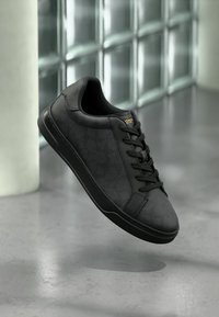 Schwarzer Ledersneaker mit Schnürsenkeln, goldenem "Coach"-Logo auf der Zunge, schwebt über einem reflektierenden Betonboden an einer Glasbausteinwand.