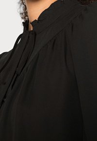 Blouse noire avec un col montant froufroutant et une fermeture par lien. Tissu doux et léger avec un plissage subtil autour des épaules.