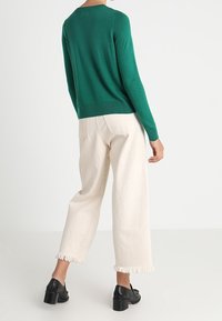 Grön stickad tröja med rundad halsringning, kombinerad med krämfärgade byxor med vida ben och fransade fållar, samt svarta klackskor.