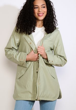 Kobieta o kręconych włosach, ubrana w jasnooliwkową kurtkę z kapturem na białej bluzce, zapinająca suwak kurtki i lekko uśmiechająca się.