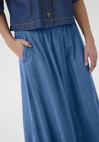 Denim nederdel med elastisk talje, der har sidelommer og et flydende design i en mellemblå nuance. Teksturen virker glat og afslappet.