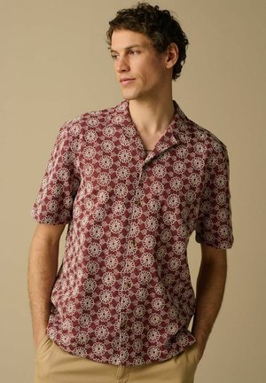 Mann trägt ein bordeauxfarbenes Kurzarmhemd mit weißen Blumenmustern, steht mit den Händen in den Taschen der beigen Hose und schaut nach links.