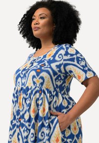 Sininen ja valkoinen kuvioitu mekko lyhyillä hihoilla, jossa on oransseja yksityiskohtia ja taskut. Kankaan pinta on pehmeä ja sileä.