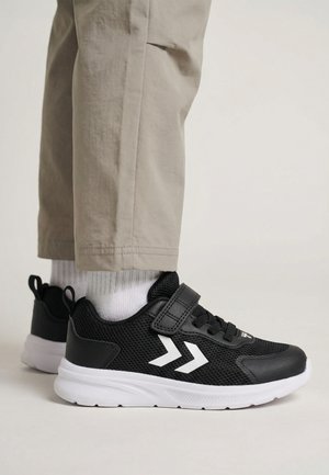 Sort-hvide atletiksko med velcrobånd, båret sammen med hvide sokker og beige bukser mod en ensfarvet lys baggrund.