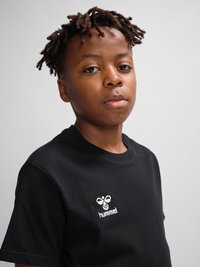 Młoda osoba z krótkimi skręconymi włosami, ubrana w czarny t-shirt hummel, patrzy bezpośrednio w kamerę na jednolitym jasnym tle.