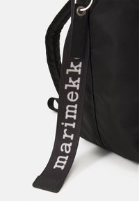 Borsa nera in tessuto testurizzato. Presenta una tracolla con l'etichetta stampata "marimekko" in bianco. Anelli tondeggianti in metallo attaccati alla borsa.