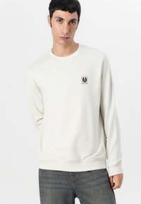 Lys beige genser med rund hals, ribbestrikkede mansjetter og et lite logo merket på brystet. Glatt stoff med et enkelt design.