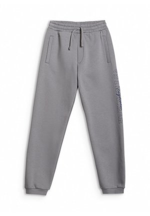 Chłopiec w czerwonej czapce typu beanie, oversize niebieskiej koszulce, szarych spodniach dresowych z napisem na nogawce oraz szarych trampkach z różowymi sznurówkami, stoi nieruchomo.