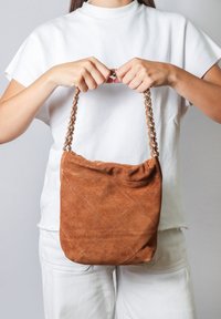 Borsa a spalla in suede marrone con motivo trapuntato, dotata di una tracolla a catena intrecciata, tenuta da una persona in camicia bianca su uno sfondo grigio chiaro.