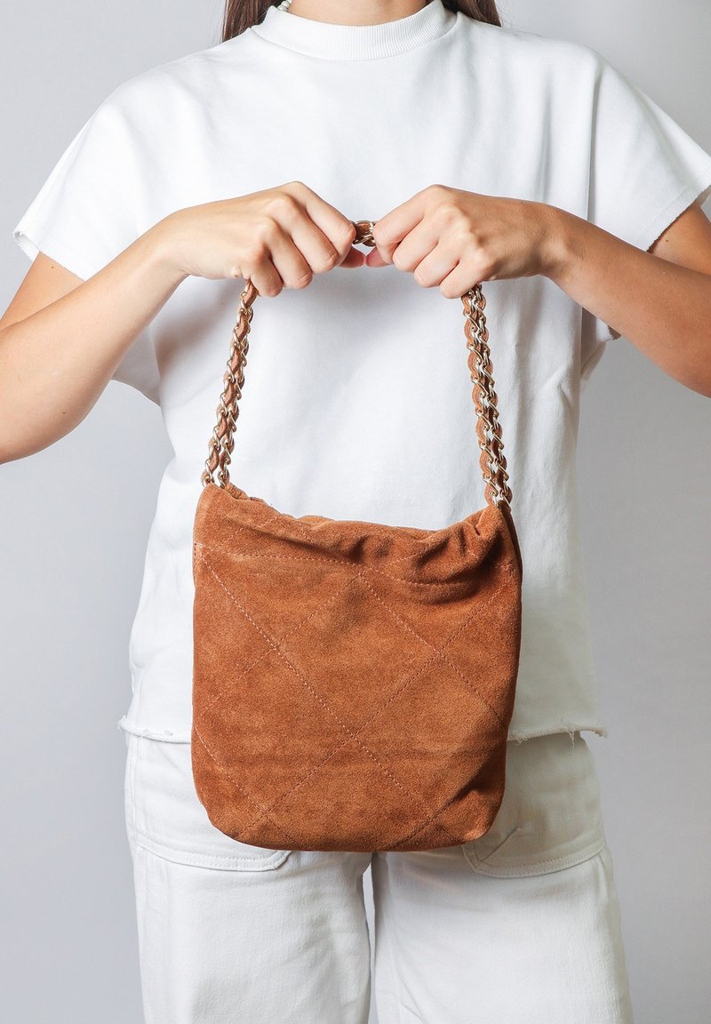 Borsa a spalla in suede marrone con motivo trapuntato, dotata di una tracolla a catena intrecciata, tenuta da una persona in camicia bianca su uno sfondo grigio chiaro.