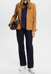 Senapsgul kavaj, marinblus, mörkblå jeans, vita sneakers. Kavajen har en enkel knapp och framfickor, slät textur.