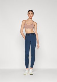 Sportovní podprsenka v taupe barvě s nastavitelnými ramínky a bezšvovým designem, doplněná o modré legíny s černým pasem a logem "NIKE PRO".