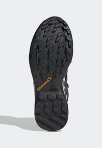 Mustat kumiset urheilukenkien pohjat, joissa on teksturoitu, kuvioitu kulutusosa. Sisältää "Continental"- ja "adidas"-brändäyksen oransseilla ja valkoisilla yksityiskohdilla.