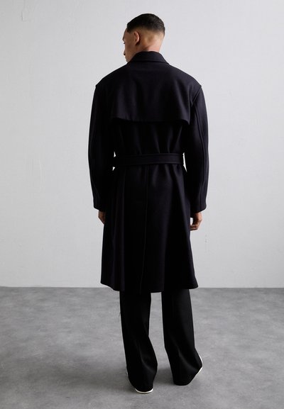 Dlouhý námořnický kabát vyrobený z texturované látky, s pásem v oblasti pasu, širokým límcem a designem zad připomínajícím plášť, kombinovaný s černými kalhotami.