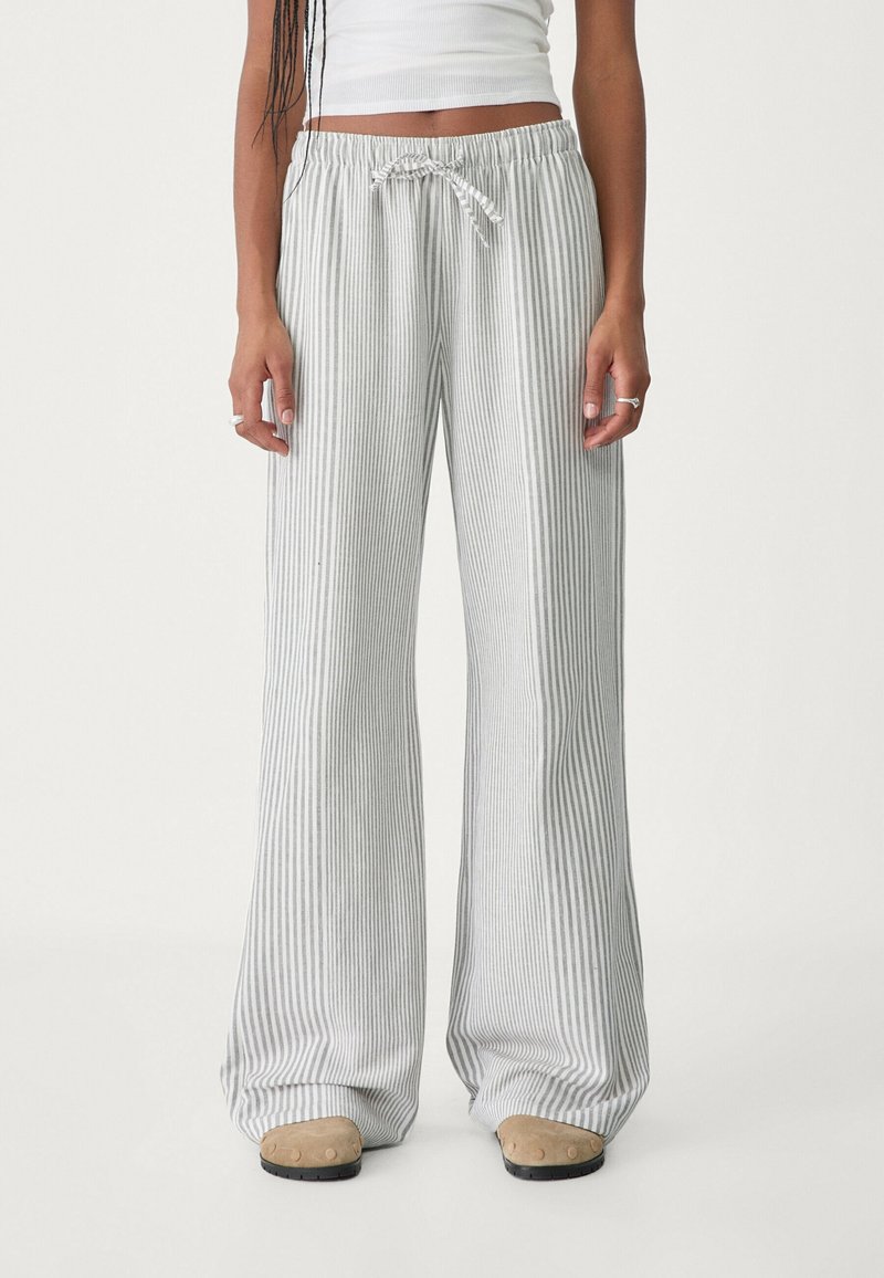 Personne portant un pantalon ample rayé verticalement gris et blanc avec une taille coulissante et des chaussures beiges, visible de la taille jusqu’aux pieds.