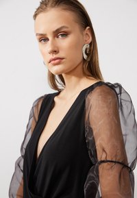 Svart kjole med dyp V-hals, prydet med gjennomsiktige, puffede ermer. Modellen har på seg store, sirkulære statementøreringer. Glatt stoff med en elegant silhuett.