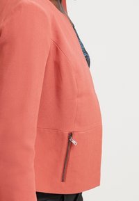 Veste couleur corail avec un design structuré, encolure arrondie et poche zippée latérale. Confectionnée en tissu texturé, silhouette ajustée.