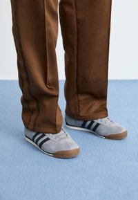 Pelēki zamša sporta apavi ar melnām svītrām, apvienoti ar brūnām treniņbiksēm ar sānu svītrām, uz zila fona.