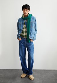 Denimjack met een lichtblauwe kleur, groen geruit overhemd eronder, donkerblauwe jeans en tan instapschoenen. Groene gebreide sjaal om de nek gewikkeld.