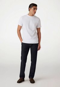 Vit crewneck-t-shirt med korta ärmar, kombinerad med mörkblåa denimjeans. Modellen bär bruna mockalofers och står mot en neutral bakgrund.