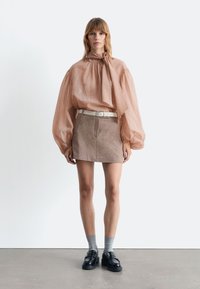 Pfirsichfarbene, leichte Bluse mit langen Ärmeln und Schleifenkragen; braurer, strukturierter Minirock; weißer Gürtel; graue Socken; schwarze Slipper.