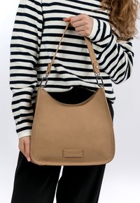 Vaahto hiekka nahkainen olkalaukku pehmeällä pinnalla, kaarevalla muodolla ja hopeisilla yksityiskohdilla. Edessä on brändilogo.