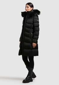Mustast pehmendusega pikk mantel, millel on eemaldatav kunstkarusnahast kapuuts, zipper-taskud ja läikiv tekstuur. Kandes musti retuuse ja saapaid.