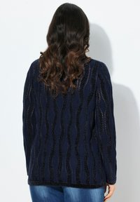 Marine cardigan met een textuur golfpatroon, lange mouwen en een open brei-design. Draag het over blauwe jeans, waarbij de achterkant zichtbaar is.