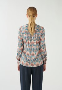 Blusa multicolore con un motivo geometrico floreale nei toni di rosa, blu e crema. Maniche lunghe e vestibilità comoda, abbinata a pantaloni a righe nere.