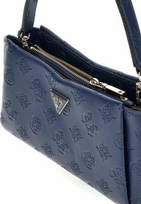 Sac à main en cuir bleu marine avec un motif de logo embossé, des accessoires en argent, une fermeture zippée, et une forme rectangulaire structurée avec une courte poignée.