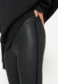 Leggings neri in ecopelle con una finitura liscia, caratterizzati da una cucitura laterale e un piccolo logo vicino alla vita.