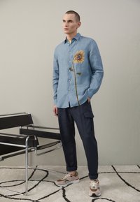 Ljusblå linneskjorta med broderad solros på vänster sida, tillsammans med marinblå cargobyxor och flerfärgade sneakers.