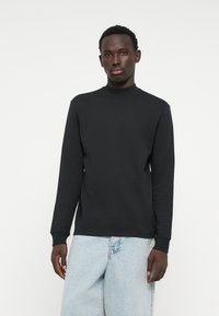 Felpa nera a maniche lunghe con collo alto, realizzata in tessuto liscio, con polsini a costine e vestibilità rilassata. Indossata con jeans azzurri chiari.