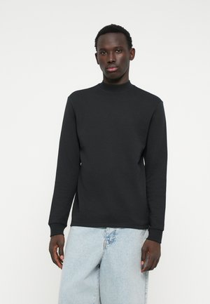 Felpa nera a maniche lunghe con collo alto, realizzata in tessuto liscio, con polsini a costine e vestibilità rilassata. Indossata con jeans azzurri chiari.