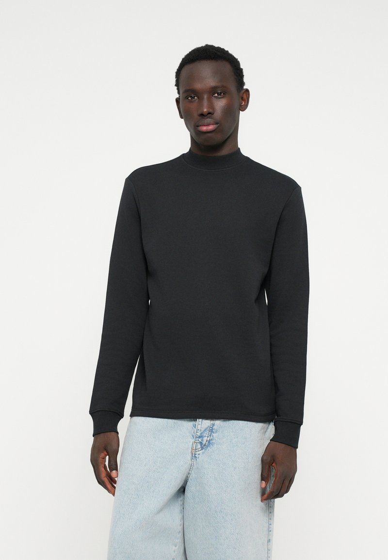 Felpa nera a maniche lunghe con collo alto, realizzata in tessuto liscio, con polsini a costine e vestibilità rilassata. Indossata con jeans azzurri chiari.