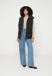 Schwarze Steppweste mit hohem Kragen, Reißverschluss und Taschen, kombiniert mit hellblauen weitem Jeans und schwarzen spitzen Ballerinas.