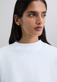 Young woman with dark hair wearing a white sweatshirt and silver hoop earring, looking to her right against a plain background.